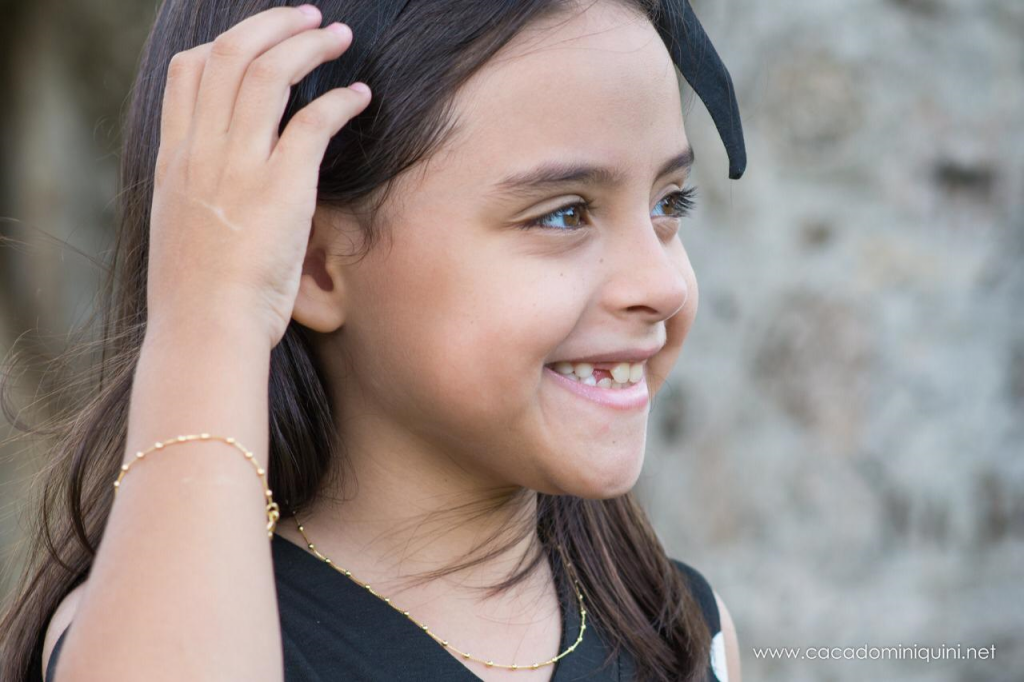 Maria Clara, 7 anos, sorrindo com a mão na cabeça usando pulseira e correntinha de bolinhas banhados a ouro 18k da marca Madonnina.